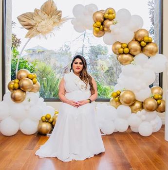 A woman in a white dress sits in front of a beautiful gold and white balloon arch for a baby shower at Seasons5